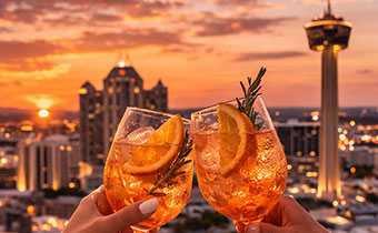 2 hands cheersing aperol spritzes with sunset in background at Tenfold